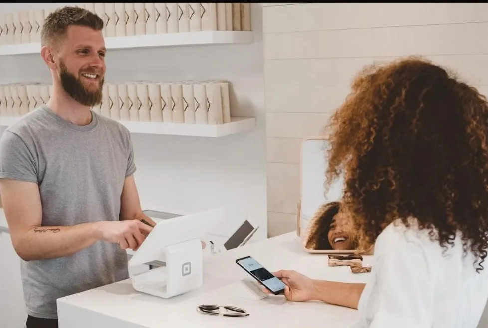 Microempreendedor sorrindo atende cliente que paga com celular aproximado à maquininha, em loja pequena